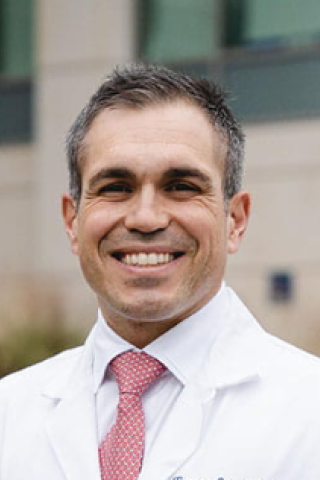 Smiling man in white lab coat and red tie outside building.