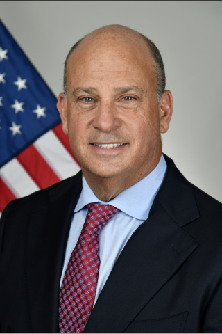 Smiling man in business attire with American flag in the background.