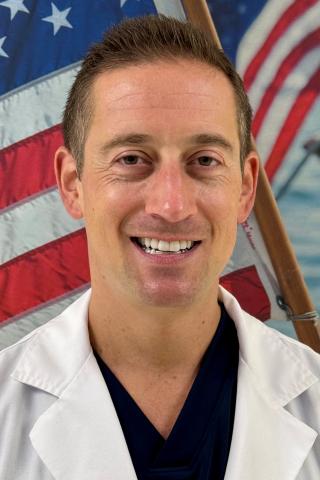 Smiling man in white lab coat with American flag in background.
