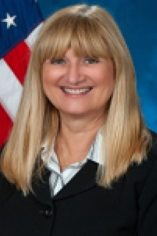 Blonde woman in a black blazer and white shirt, smiling in front of an American flag.