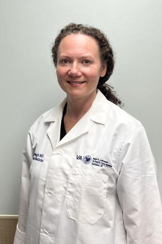 A smiling woman in a white lab coat stands against a beige wall.