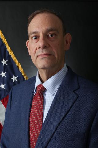 Man in suit and red tie stands in front of American flag.
