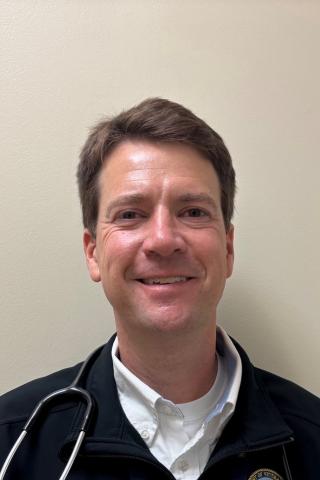 Man with short hair and stethoscope smiling against a light-colored wall.