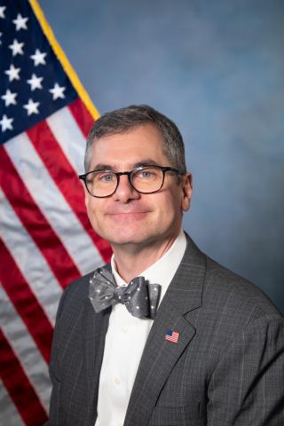 Man in gray suit and bow tie stands in front of American flag.