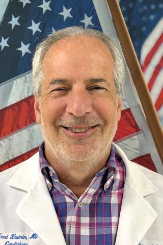 Smiling man in white lab coat and plaid shirt with American flag in background.