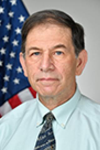 A man in a light blue shirt and tie stands in front of an American flag.