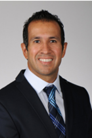 Man in black suit and blue tie smiling against a gray background.