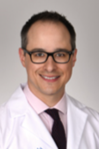 A smiling man in a white lab coat, pink shirt, and black tie.