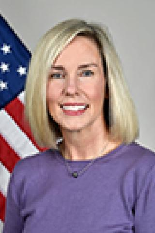 Portrait of a woman with blonde hair, smiling, and the American flag in the background.