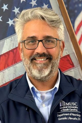 A smiling man with a beard and glasses in front of an American flag.