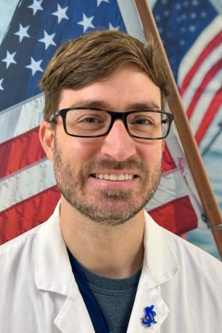 Man in white lab coat and glasses smiling in front of an American flag.