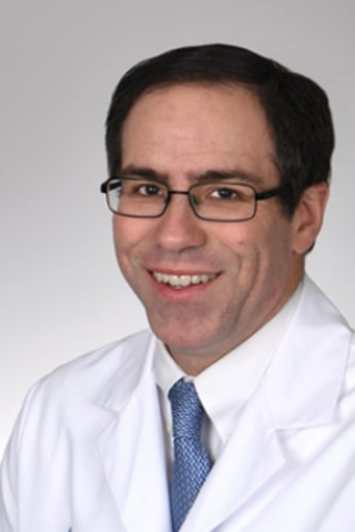 Smiling man in white lab coat and blue tie, wearing glasses.