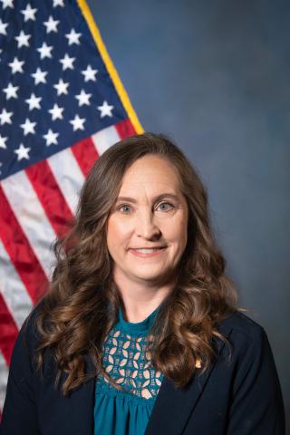 Woman with long brown hair and blue blouse, smiling in front of an American flag.