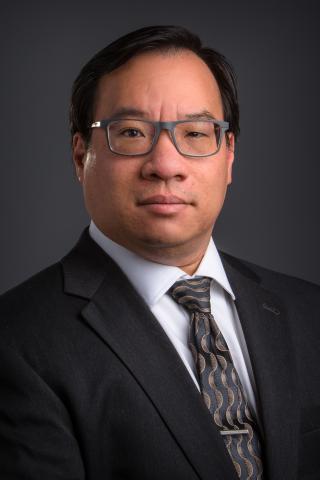 Man in black suit and tie, wearing glasses, against a black background.