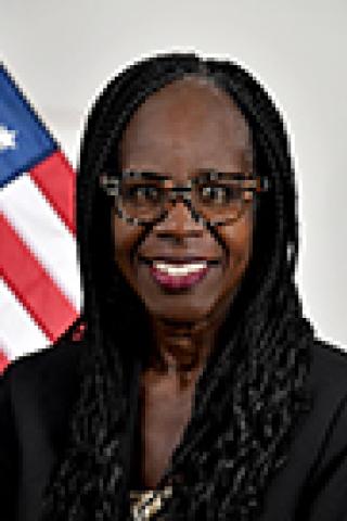 Woman in black blazer and glasses smiling in front of American flag.
