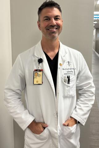 Smiling male doctor in white lab coat with name tag stands in hallway with hands in pockets.