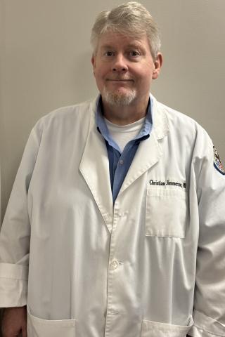 Man in white lab coat with blue shirt underneath, smiling against a plain wall.