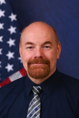 A man with a beard wearing a tie and shirt, in front of the US flag.
