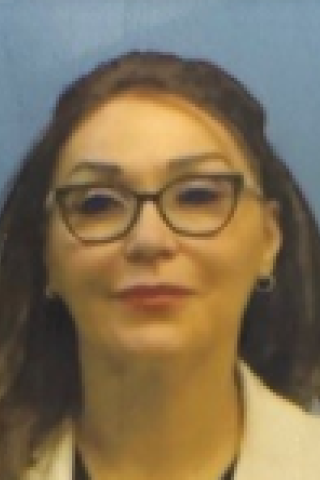 A woman with long hair, wearing glasses and a white jacket, smiling for the camera.