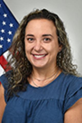 Woman with curly hair wearing blue blouse smiling in front of American flag.