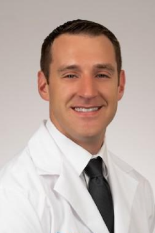 Male doctor in white lab coat and black tie smiling at camera.