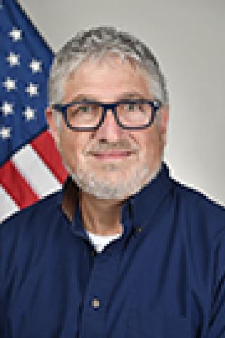 Man with gray hair, glasses, and blue shirt standing in front of an American flag.