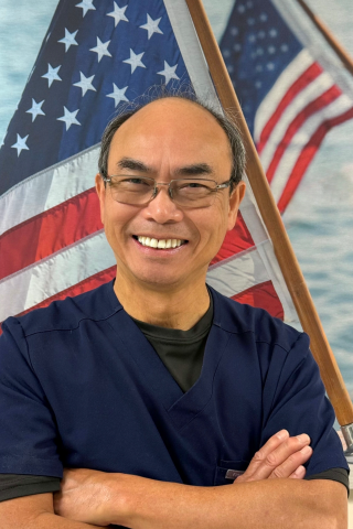 Man in blue shirt smiling with arms crossed in front of American flag.
