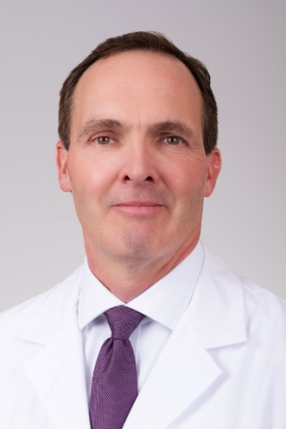 A man in a white lab coat and purple tie smiles for the camera.