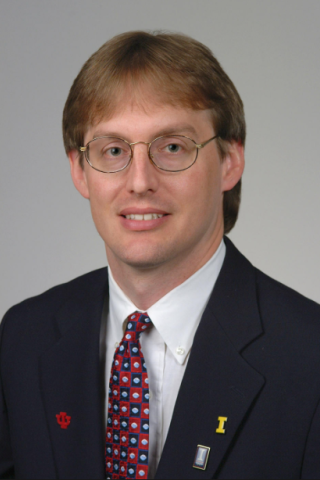 A man with glasses and a blue suit with a red and blue tie smiles.