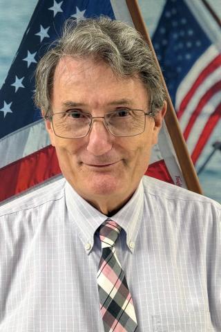 Man with gray hair, glasses, and plaid tie stands before two American flags.