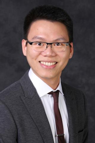 A smiling man in a gray suit and brown tie, wearing glasses.