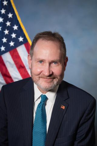 Smiling man in suit and blue tie stands in front of American flag.