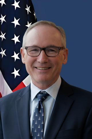 Portrait of a man in suit and tie, smiling against an American flag.