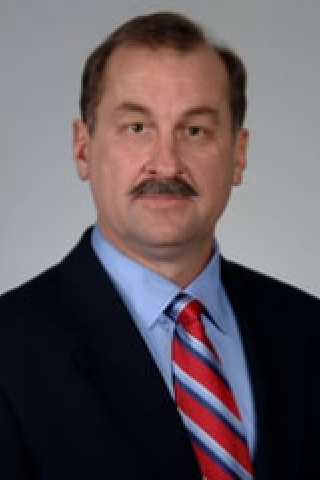Man with mustache in black suit and blue shirt with red and white striped tie.