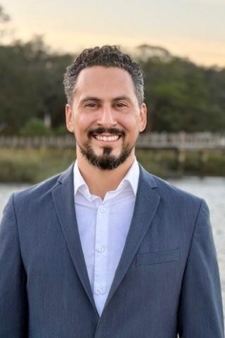 Man in a blue suit and white shirt smiles by a waterfront.