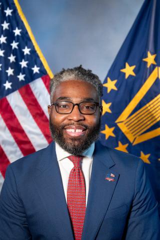 Man in formal suit and red tie smiling in front of American and Indiana flags.