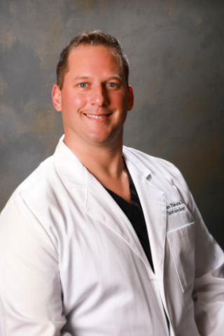 Smiling man in white lab coat and black shirt poses against gray background.