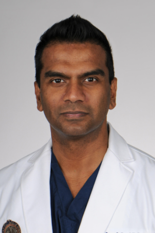 A man in a white lab coat and blue shirt stands against a white background.