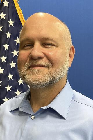 Man with a bald head and beard smiles in front of an American flag.