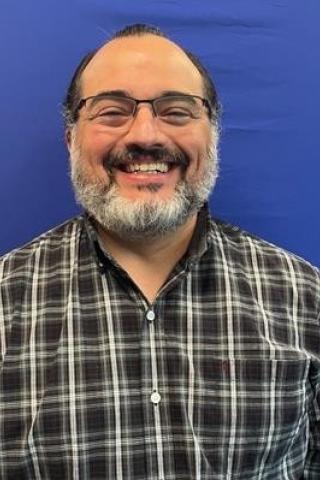 A man with glasses and a beard smiles against a blue backdrop.