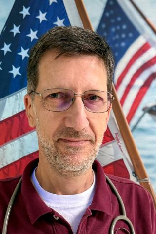 A man with a beard and glasses wearing a maroon shirt and stethoscope in front of an American flag.