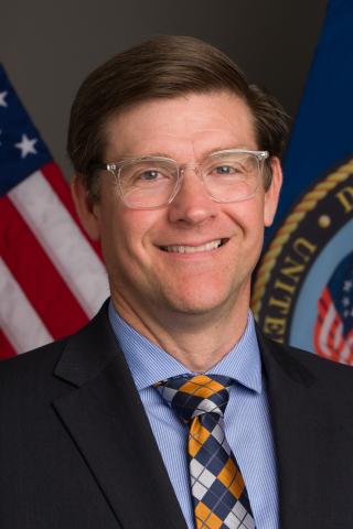 A white male smiles for his official portrait with a U.S. flag and VA flag in background