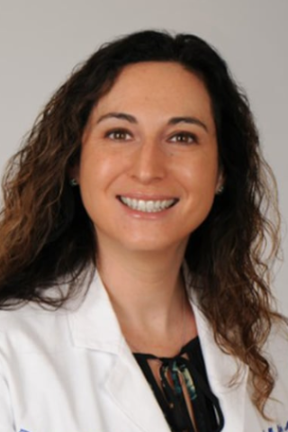 A smiling woman with curly hair in a white lab coat and black inner shirt.