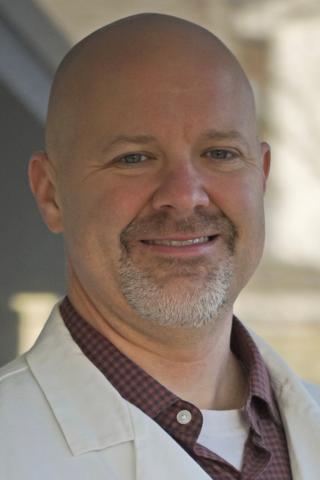 Photograph of a smiling bald man in a white coat and red shirt.