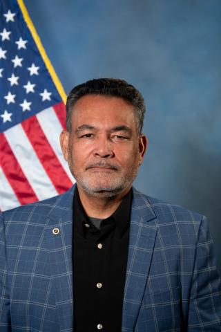 Man in blue blazer and black shirt standing in front of American flag.