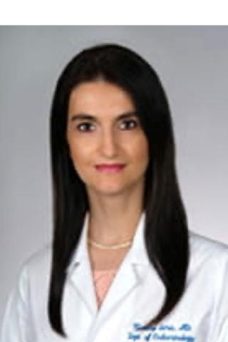 A smiling woman with long black hair wearing a white lab coat and necklace.