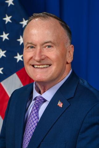 Man in suit and purple tie smiling with American flag in the background.