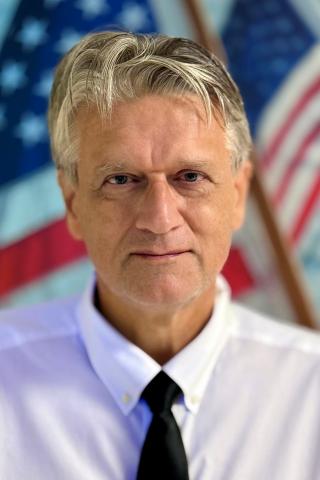 Portrait of a man in a white shirt and black tie, standing in front of an American flag.