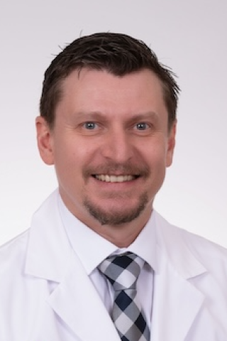 A smiling man in a white lab coat and checkered tie.