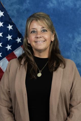Woman in black top and beige cardigan smiling in front of American flag.